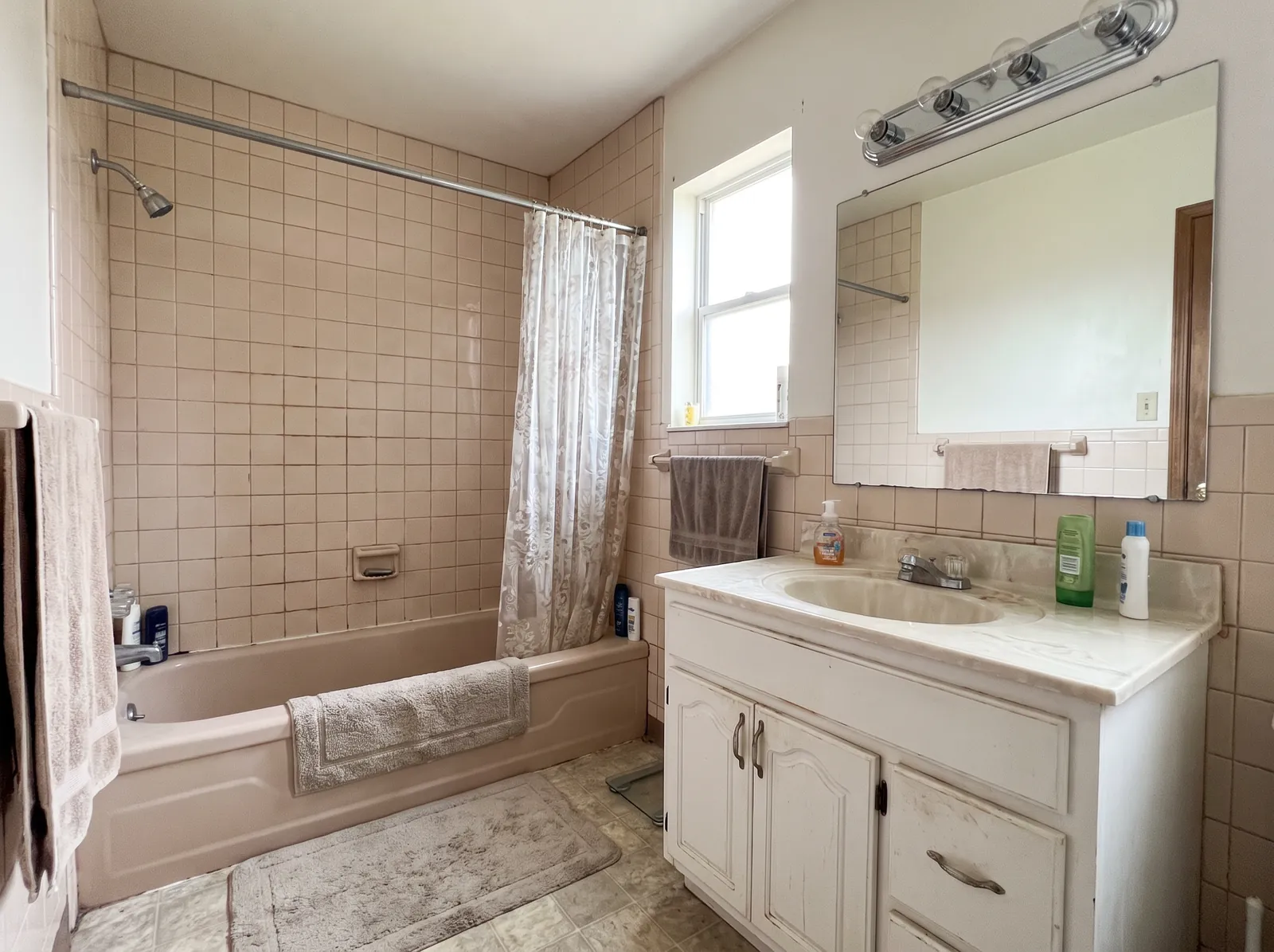 Before: a small bathroom with a pink tub, small pink-beige wall tiles and a white vanity under a bar light